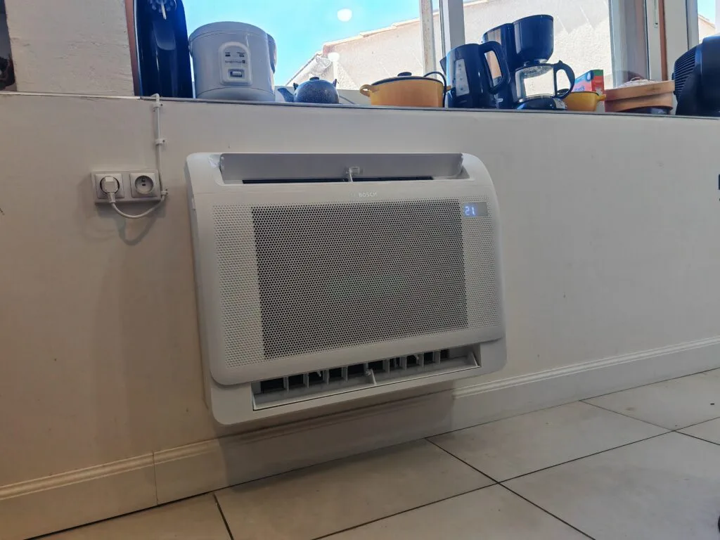 Console de climatisation réversible posée au bas d’un mur de cuisine, sous la fenêtre, affichant 21 degrés