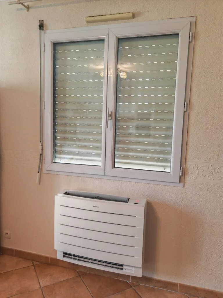 Console de pompe à chaleur air-air posée au sol sous une fenêtre, dans un séjour carrelé