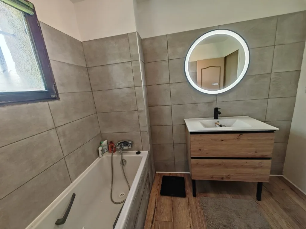 Salle de bains rénovée : grands carreaux gris, meuble vasque en bois et miroir rond lumineux