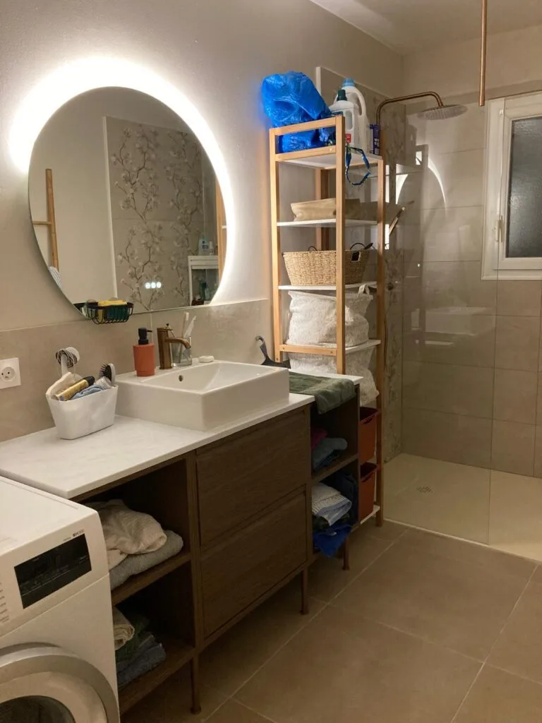 Salle de bains rénovée avec miroir rond rétroéclairé, vasque à poser sur meuble bois et douche à l’italienne